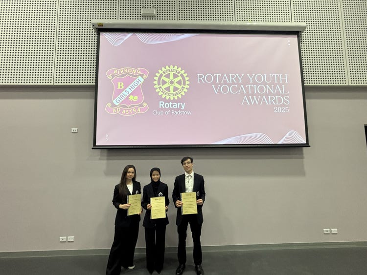 Three students stand holding Rotary Youth Vocational Awards on stage beneath a large projector that displays Rotary Youth Vocational Awards