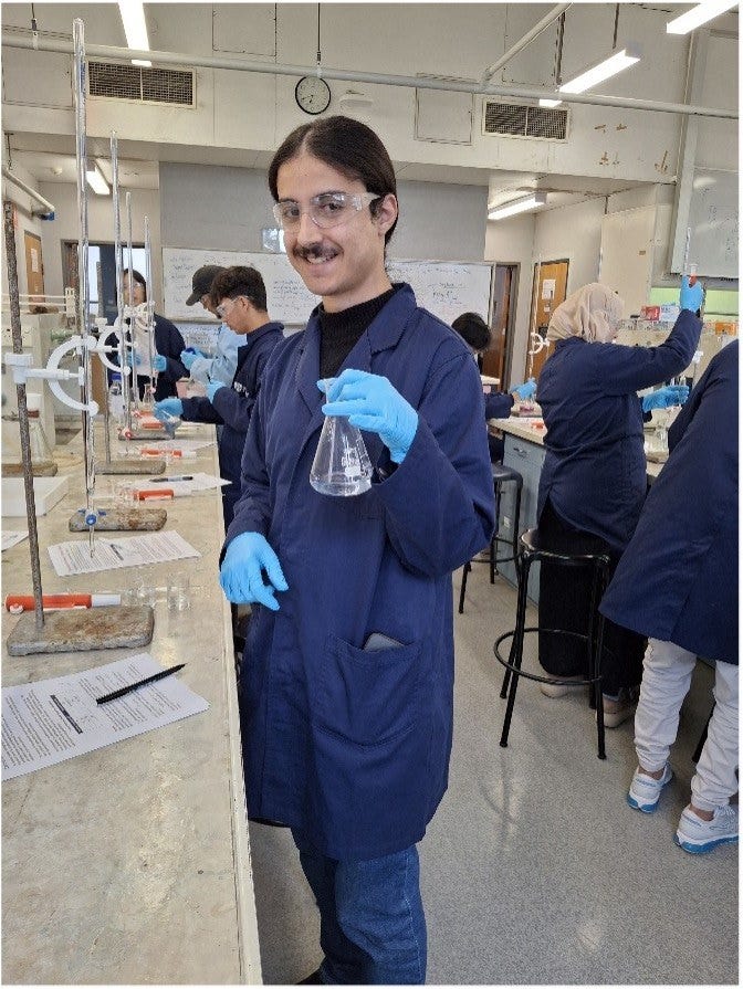 Student stands in a laboratory wearing protective clothing, goggles and holds a beaker