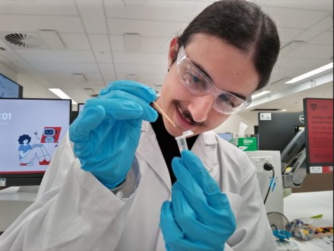 Student dressed in lab gown, wearing goggles and gloves deposits liquid into a beaker in a science lab
